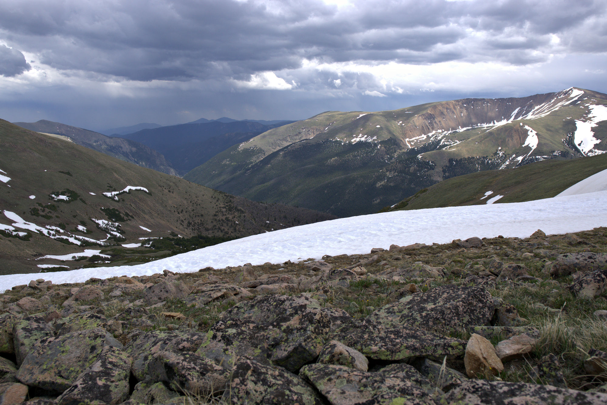A distant mountain range, viewed from the top of a mountain. Dark clouds gather above the mountain range and the sky is grey.